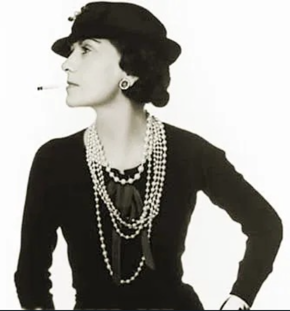 Coco Chanel em pose icônica, vestindo colares de pérolas em várias voltas.