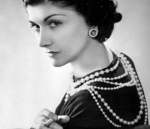 Coco Chanel vestindo vários colares de pérolas, com um olhar icônico e sofisticado.