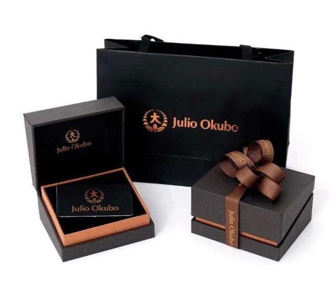 Embalagens de presente da marca Julio Okubo, incluindo caixas e sacola com o logotipo da marca em dourado.