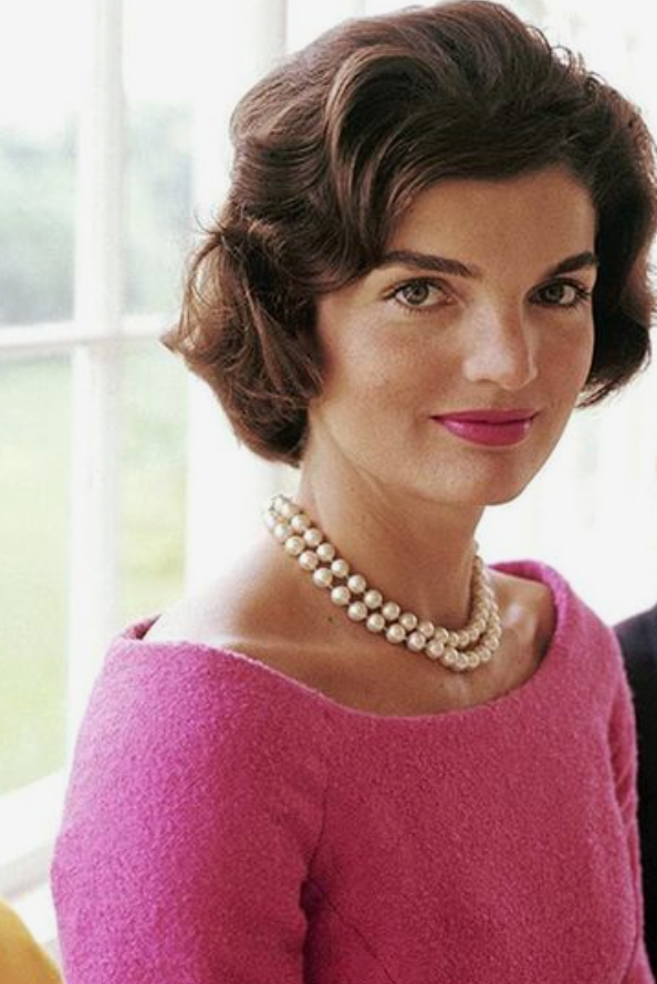 Jacqueline Kennedy Onassis usando um colar de pérolas clássico, combinando com um elegante vestido rosa.