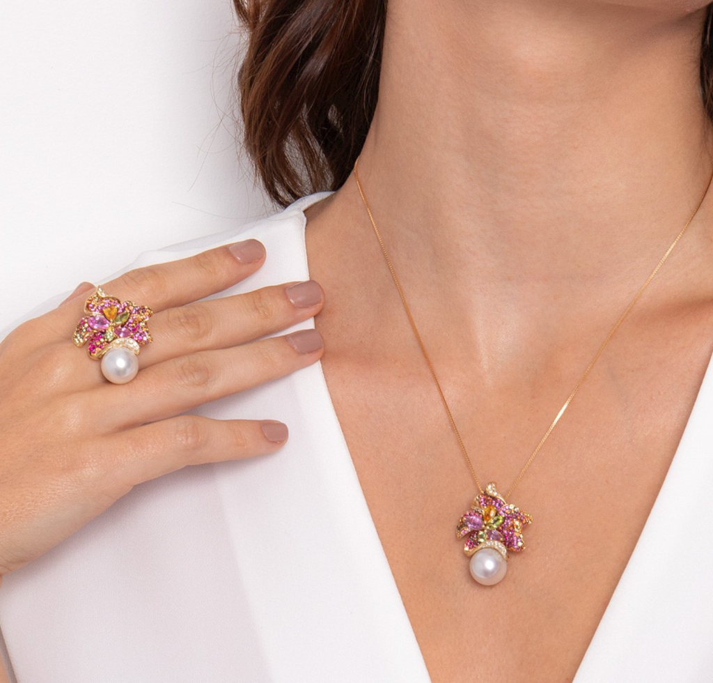 Mulher usando joias com pingente e anel de flor em ouro rosé, adornados com pérolas e pedras coloridas.